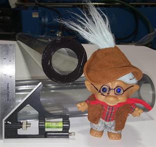 A troll doll and ruler with level next to two heavy walled tubes crossing each other.