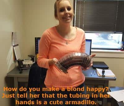 A employee holding a mitered bend. Caption says, "How do you make a blond happy? Just tell her that the tubing in her hands is a cute armadillo."