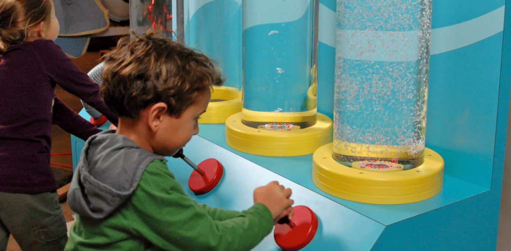 Children using an interactive display to shoot water and confetti into clear plastic tubes.